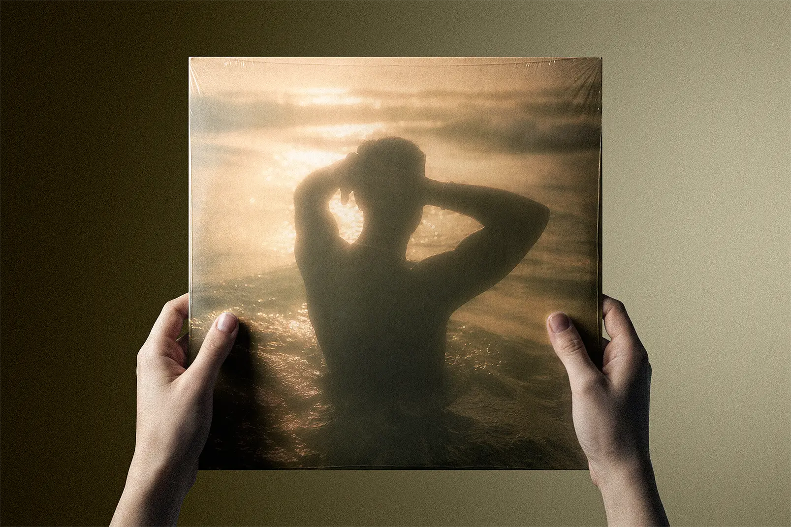 Two hands holding a square cover artwork of a silhouetted figure with arms raised, standing in the sea at sunset.