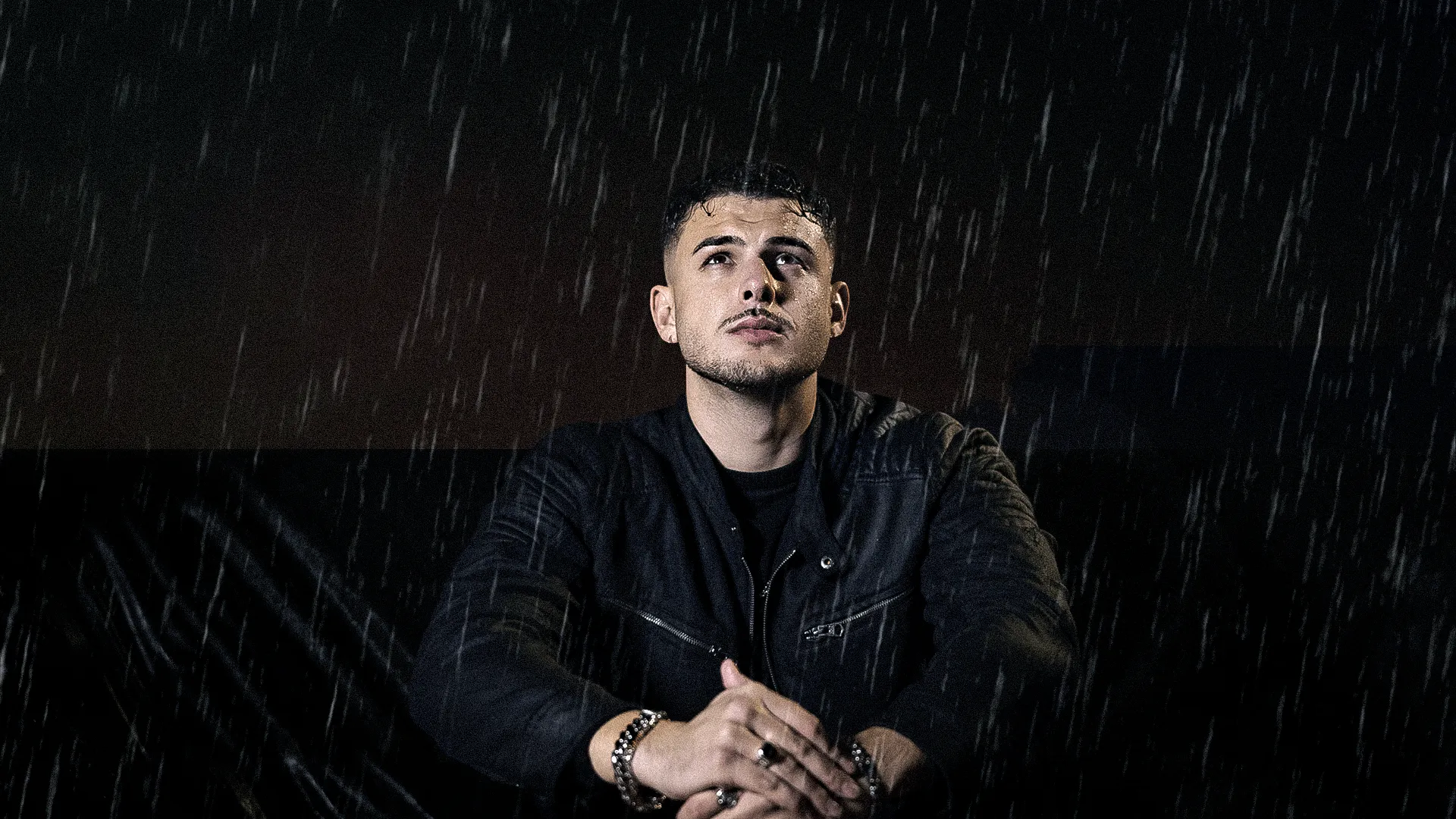 Cinematic portrait of a male artist sitting in the rain at night, wearing a black jacket and looking upward in a dramatic dark setting.