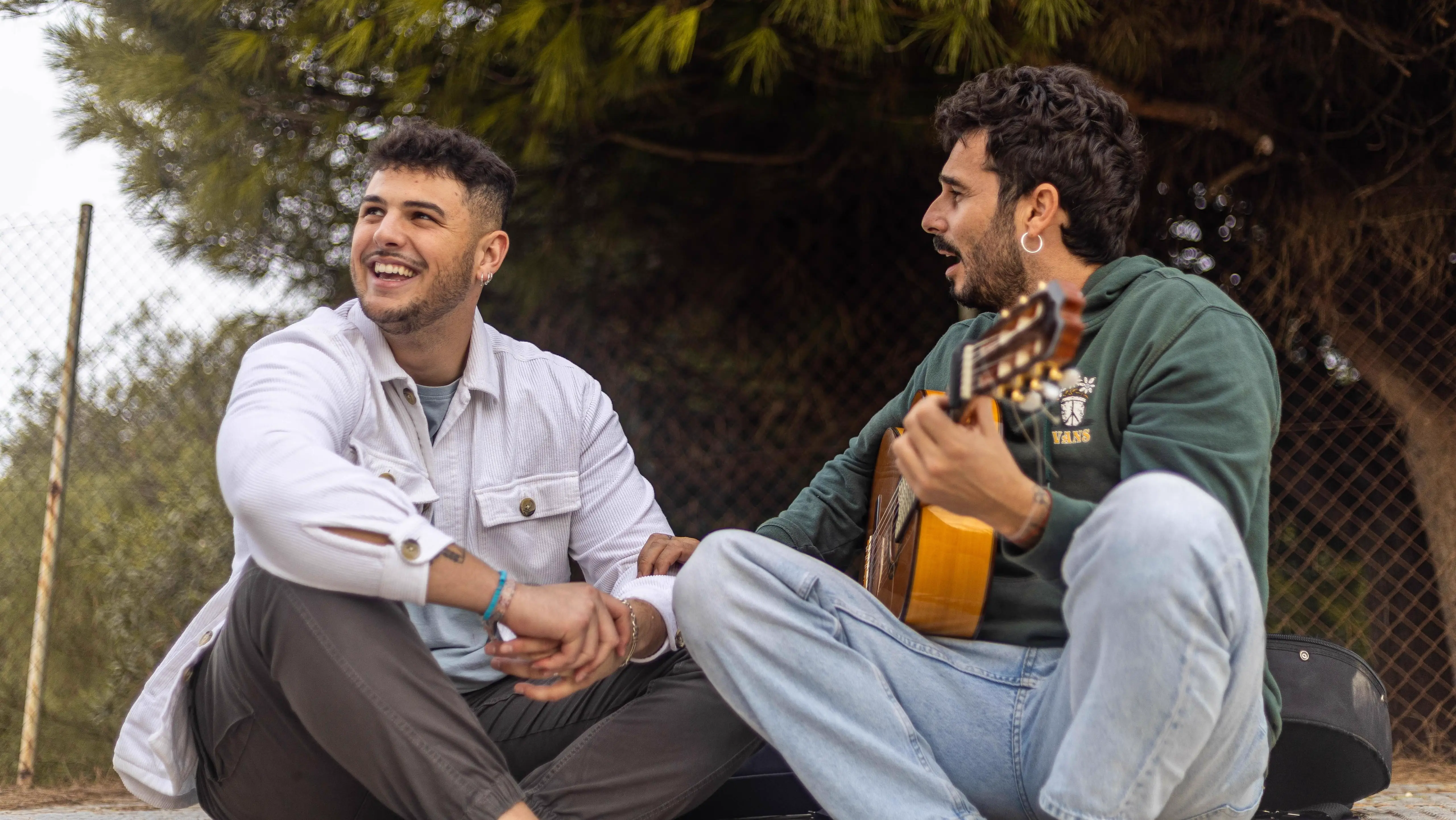 Two artists sitting outdoors in a relaxed setting, smiling and talking while one holds a Spanish guitar