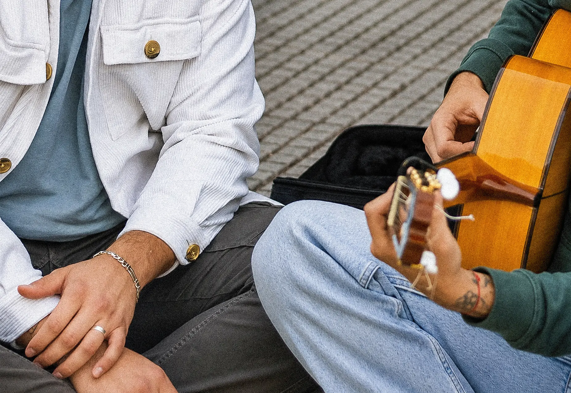 Close-up detail of two artists sitting during an acoustic session, with a Spanish guitar being played in an outdoor setting.